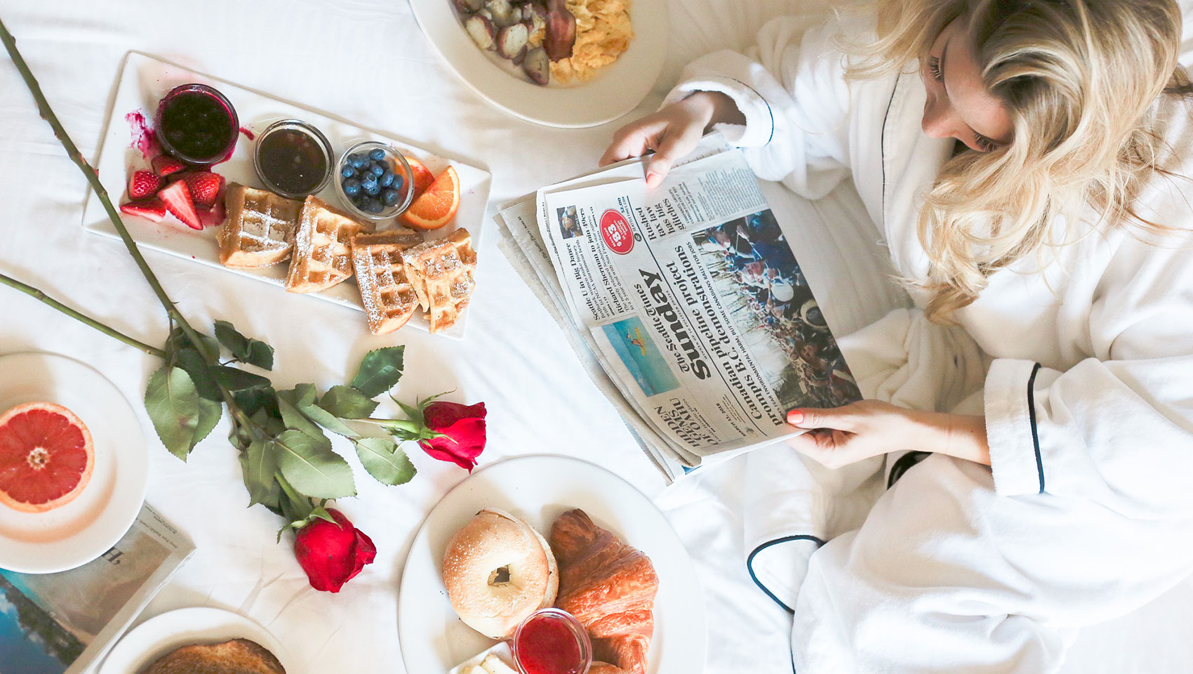 Breakfast spread on bed with roses, newspaper and blonde woman in white robe